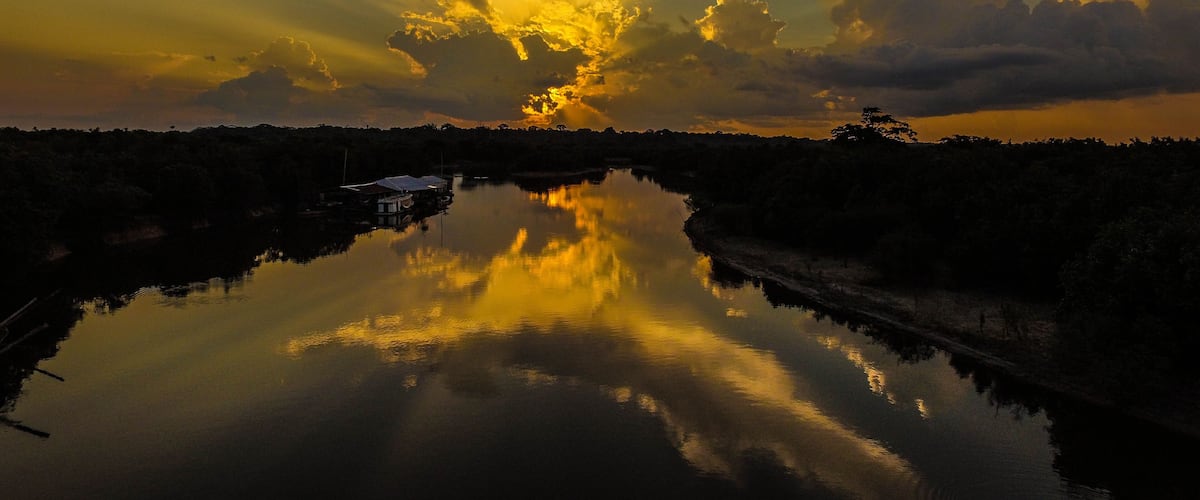 A sunset over a calm river with a house on the left bank and dense trees in the background.