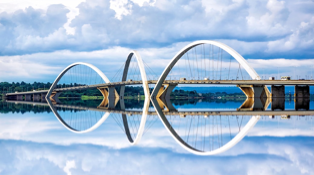 A modern bridge with a unique arch design spanning over a calm body of water, reflecting the structure and sky.
