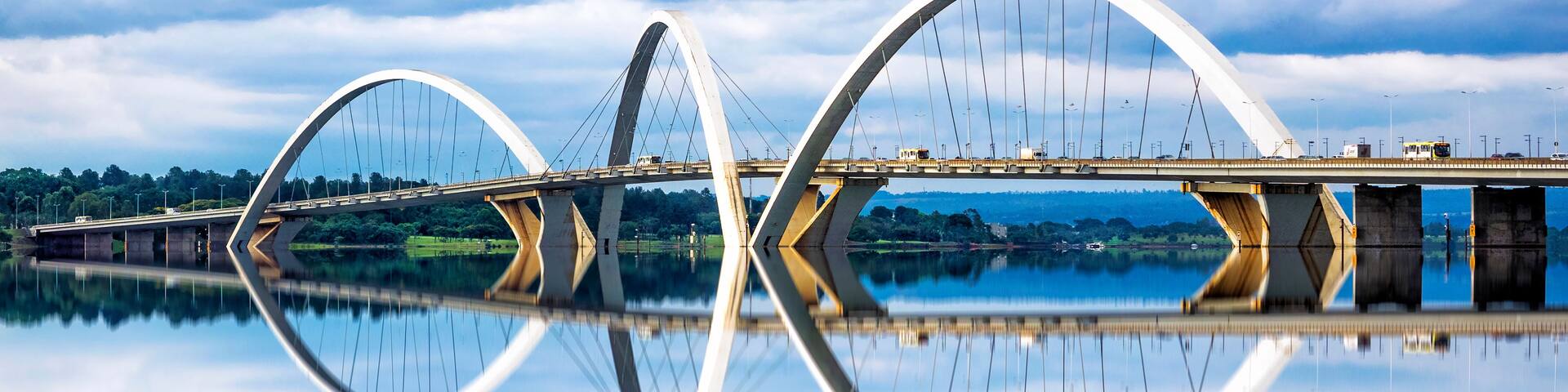 A modern bridge with a unique arch design spanning over a calm body of water, reflecting the structure and sky.