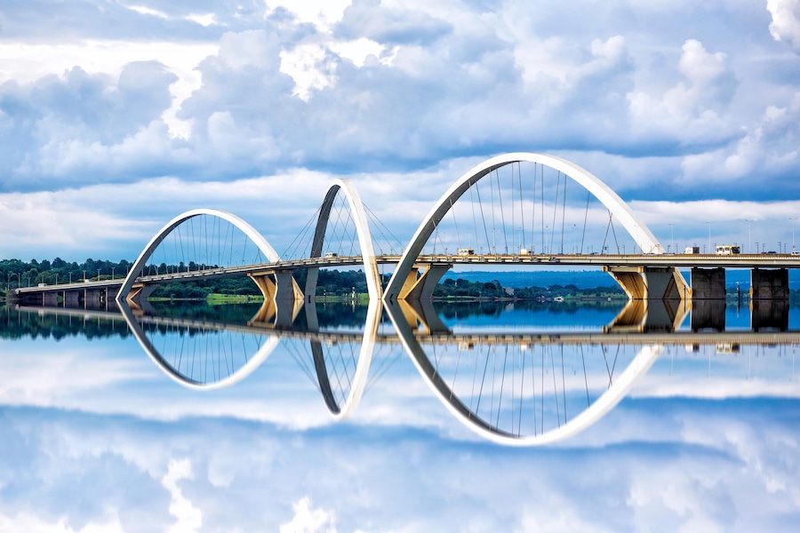 A modern bridge with a unique arch design spanning over a calm body of water, reflecting the structure and sky.