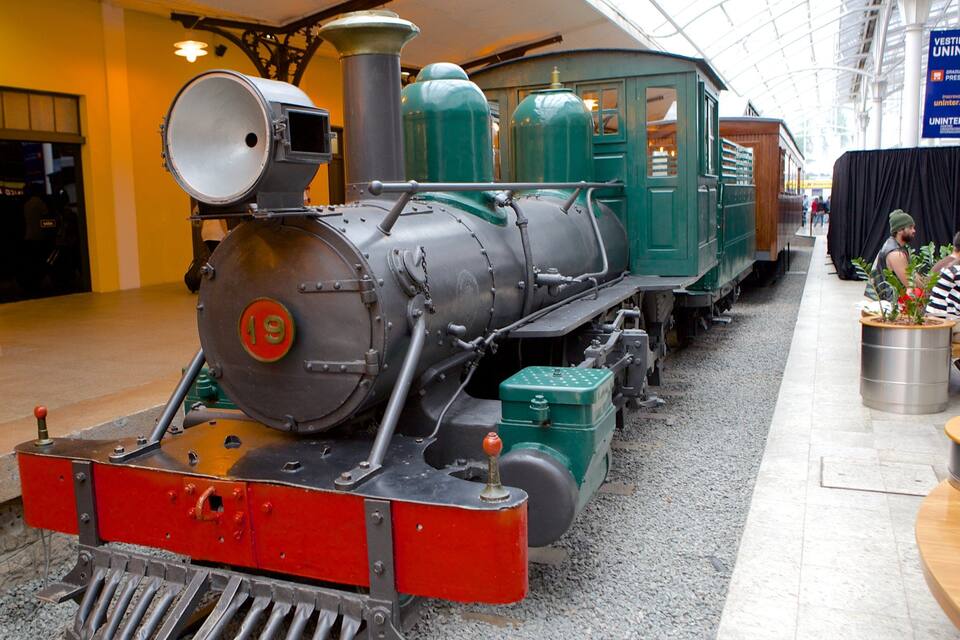 A vintage steam locomotive with a red front and green body, displayed indoors with a glass ceiling above.