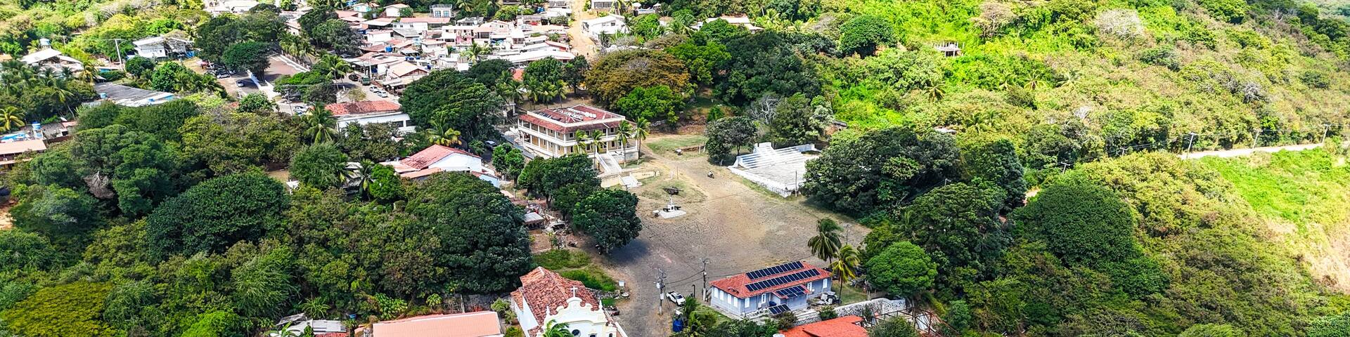 Vista aérea da Vila dos Remédios em Fernando de Noronha, com casas coloridas, igrejas históricas e vegetação tropical cercadas por mar azul, refletindo o charme e a cultura local