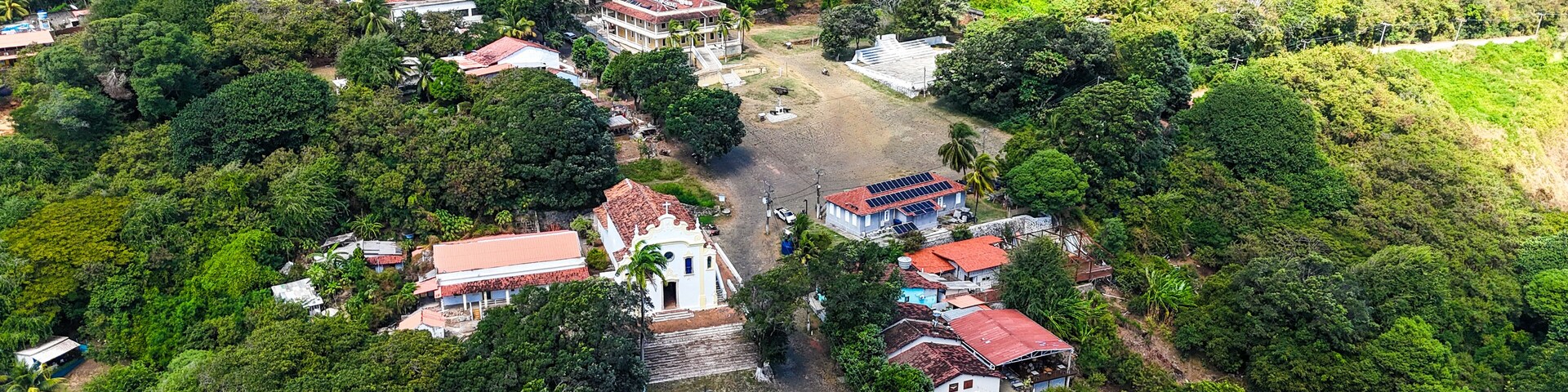 Vista aérea da Vila dos Remédios em Fernando de Noronha, com casas coloridas, igrejas históricas e vegetação tropical cercadas por mar azul, refletindo o charme e a cultura local