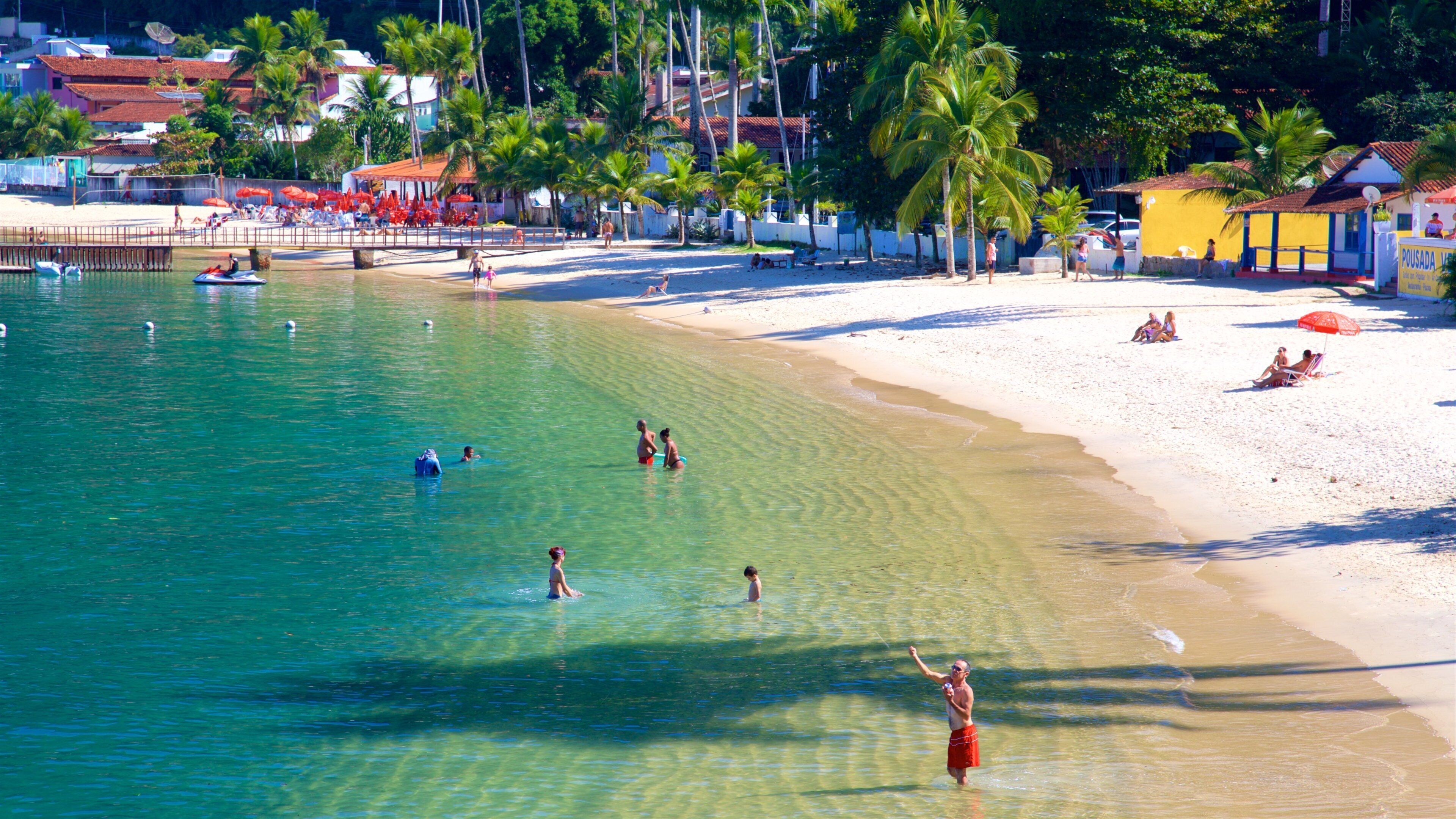 A beach with clear turquoise water, people swimming and sunbathing, palm trees, and buildings in the background.