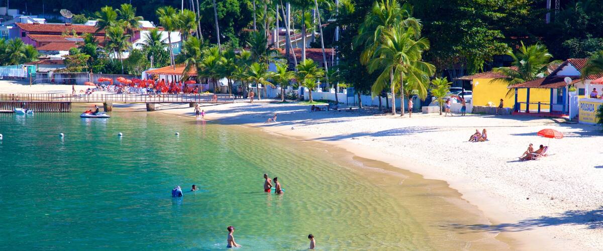 A beach with clear turquoise water, people swimming and sunbathing, palm trees, and buildings in the background.