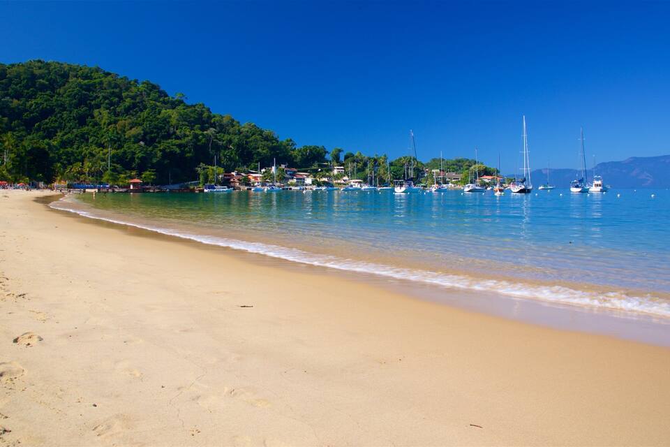 A sandy beach with clear blue water, boats, and a forested hill in the background.