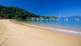Une plage de sable fin bordée d’une eau d’un bleu limpide, avec des bateaux et une colline boisée en arrière-plan.