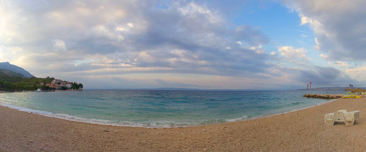 A pebbled beach with clear turquoise water, a small hut, and a mountain in the background.