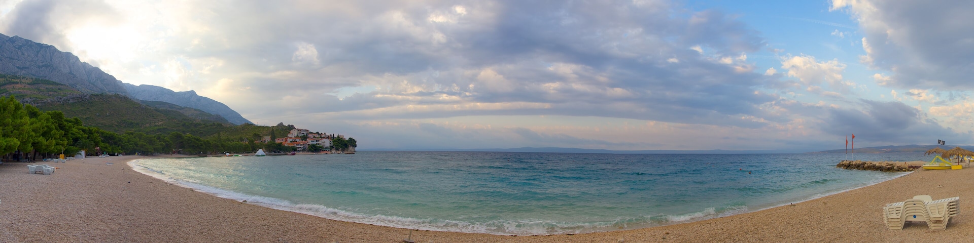 A pebbled beach with clear turquoise water, a small hut, and a mountain in the background.
