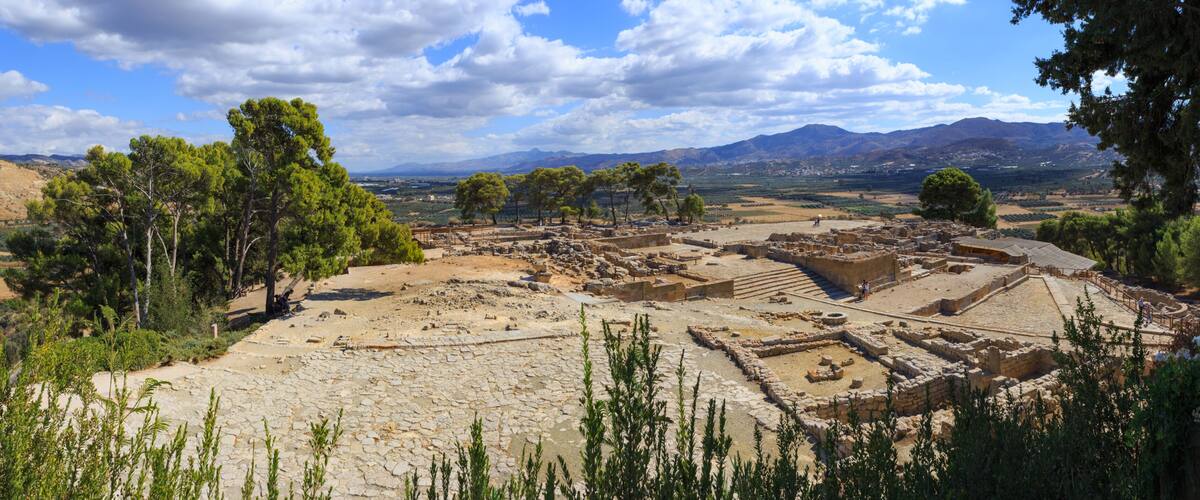 Ancient ruins with stone pathways and structures, surrounded by trees and mountains in the background.