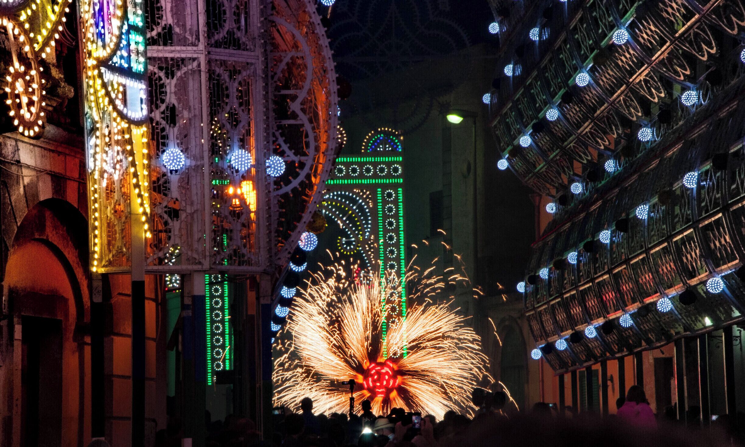A nighttime street with illuminated buildings, a Ferris wheel, and fireworks.