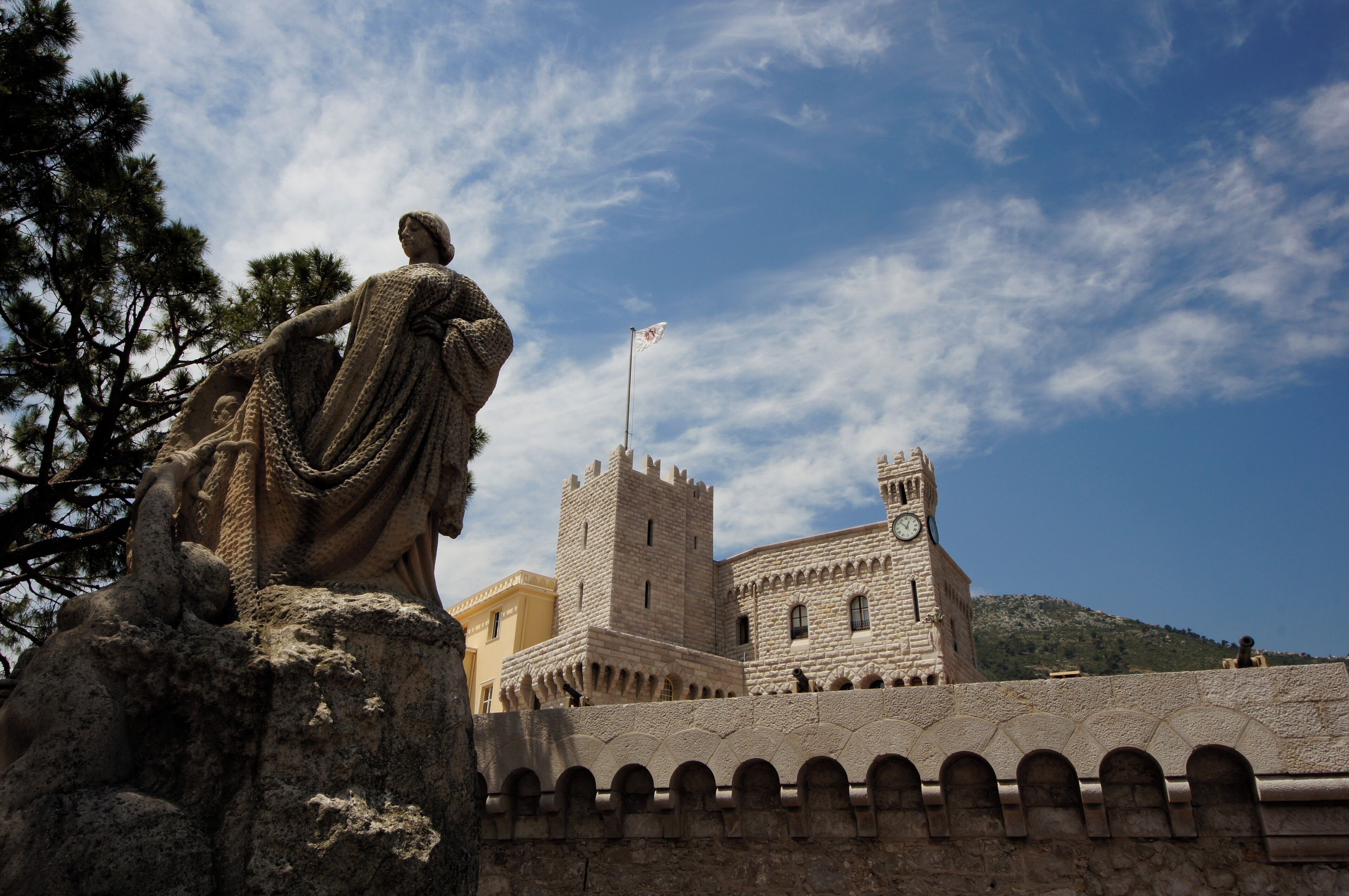 A stone statue of a woman in a flowing robe, a castle with a clock tower, and a clear blue sky with clouds.