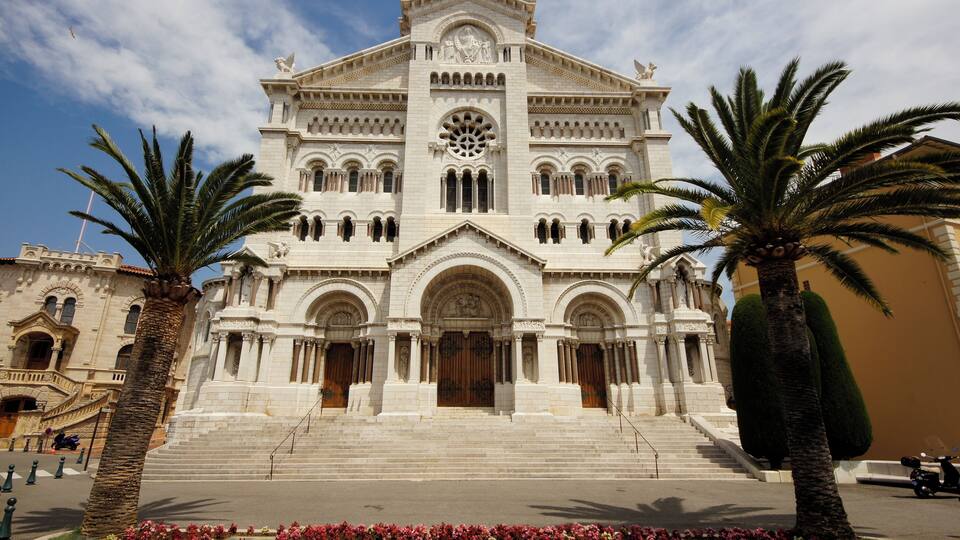 A grand white building with arched windows and a central circular window, flanked by palm trees and a flower bed.