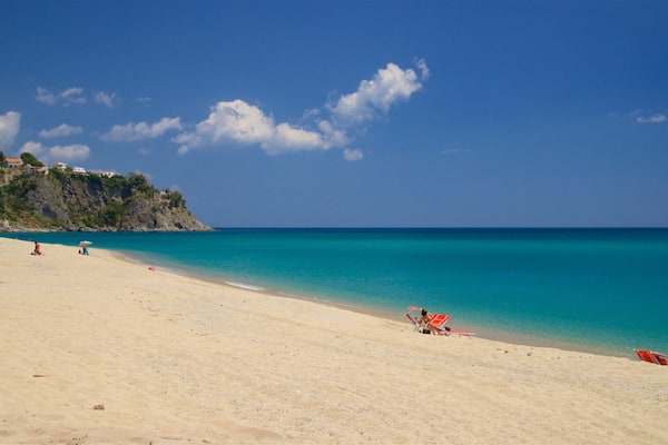 A beach with clear turquoise water, a sandy shore, and a rocky cliff with buildings on top.