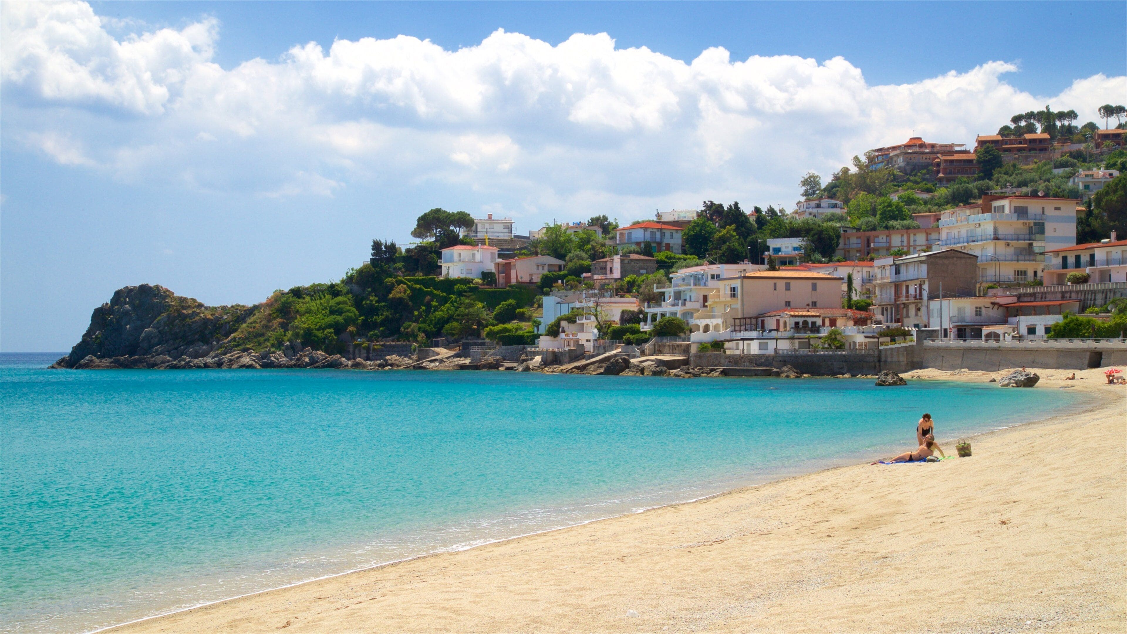 A beach with clear turquoise water, a sandy shore, and a hillside with colorful buildings.