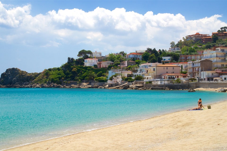 A beach with clear turquoise water, a sandy shore, and a hillside with colorful buildings.