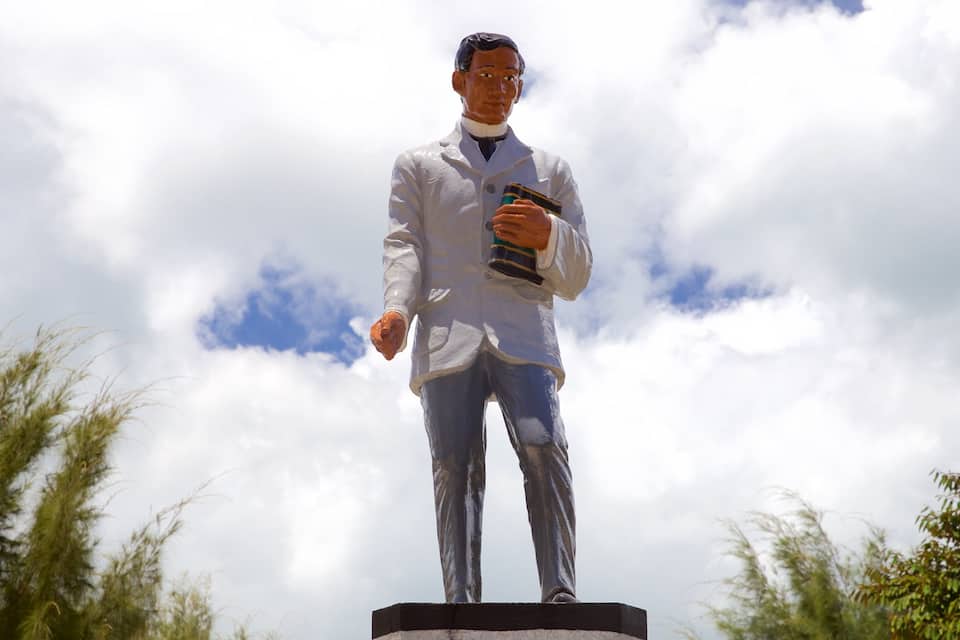 Eine Statue eines Mannes in einem weißen Kittel, der ein Buch hält, auf einem Sockel, im Hintergrund Bäume.