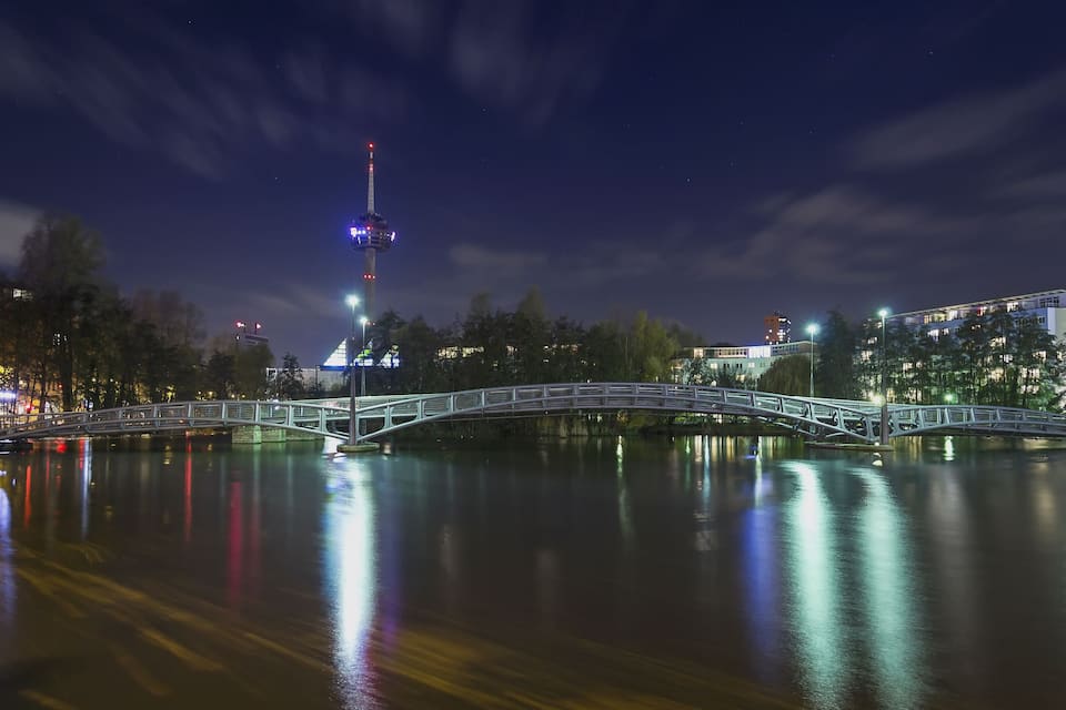 Nachtszene mit einer Brücke über einem ruhigen Fluss, Stadtlichtern und einem Turm mit Beleuchtung.