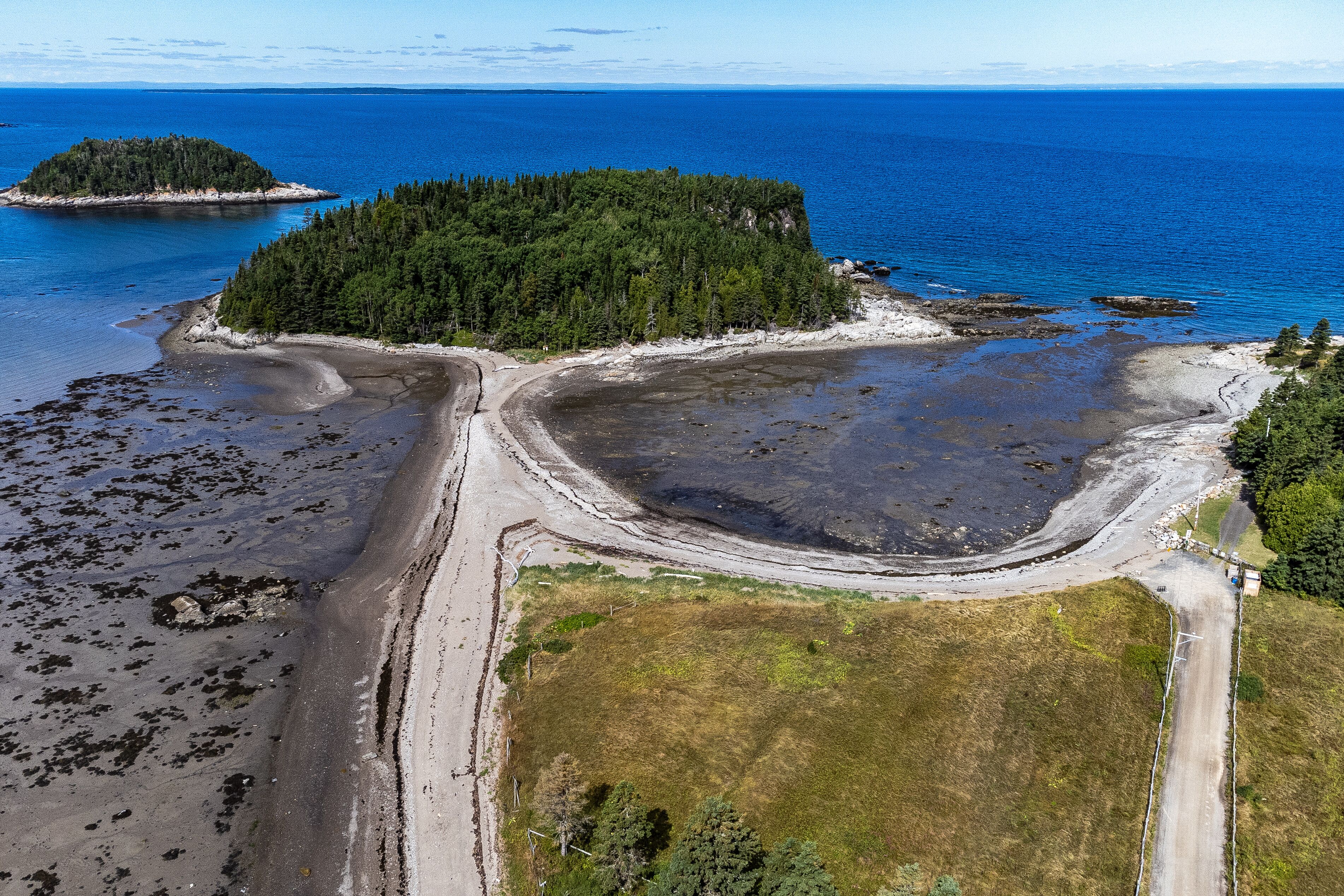 An aerial view of a coastal area with a forested island, a sandy beach, and a clear blue sea.
