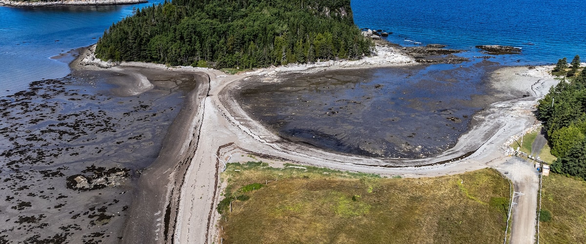 An aerial view of a coastal area with a forested island, a sandy beach, and a clear blue sea.