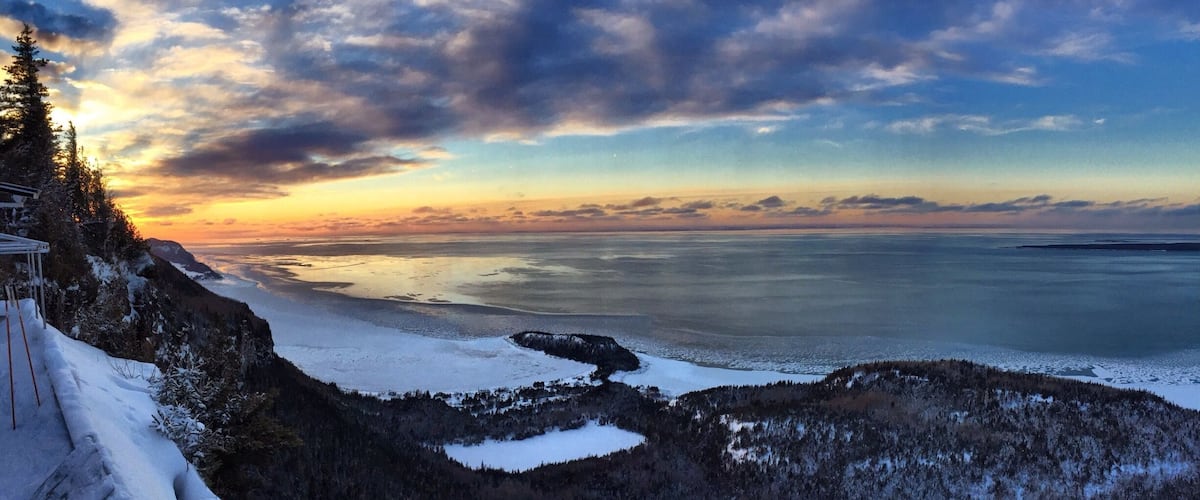 A snowy landscape with a frozen lake and a forested mountain range during sunset.