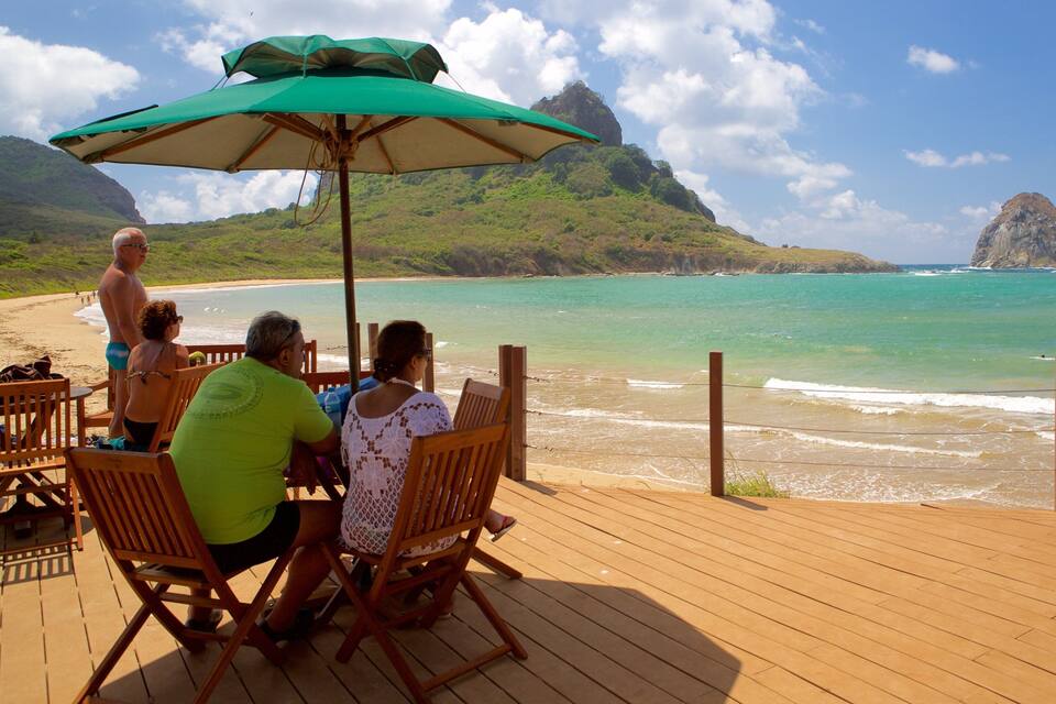 A wooden deck overlooking a beach with clear turquoise water, a green umbrella, and a mountainous landscape in the background.