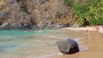A tropical beach with clear turquoise waters, a large rock in the foreground, and people engaging in water activities.