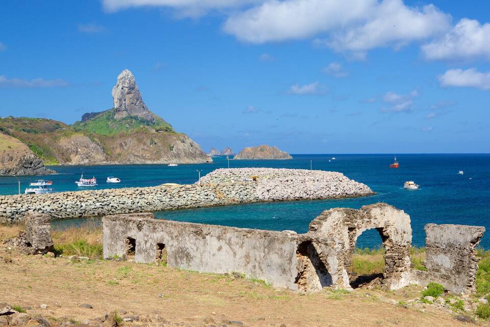 A coastal landscape with a prominent rocky formation, a stone wall, and boats on the water.