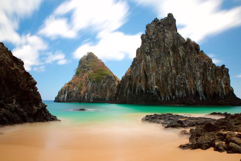 A coastal landscape with two prominent rock formations, a sandy beach, and turquoise waters.