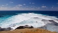 Un paisaje costero con olas rompiendo contra rocas y un cielo azul despejado.