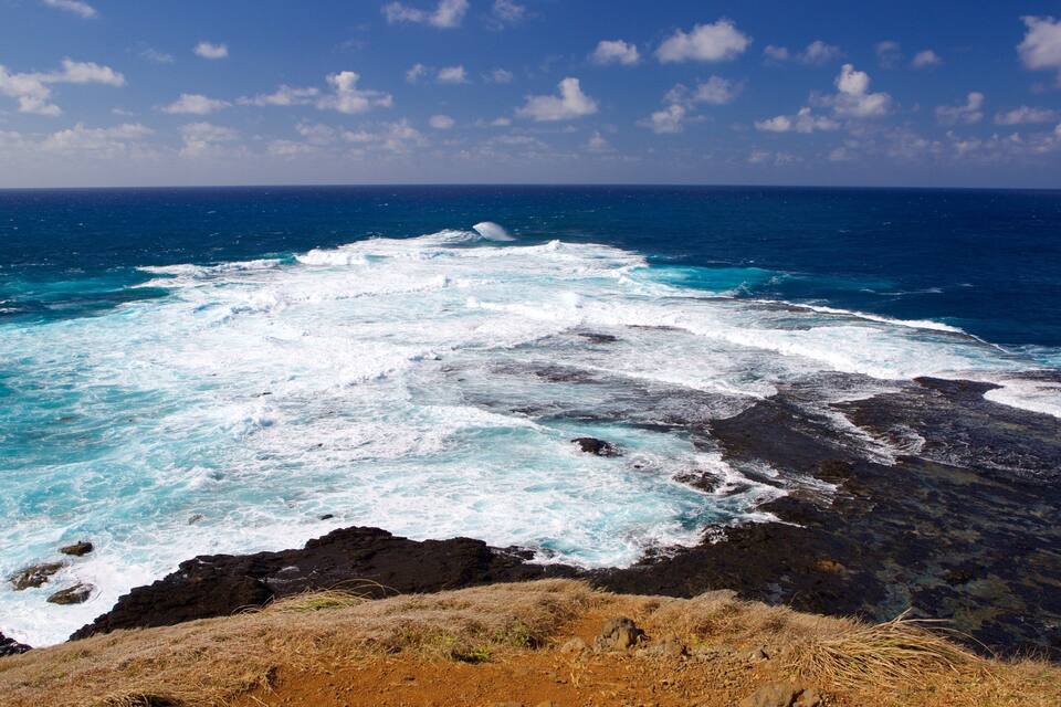 A coastal landscape with waves crashing against dark rocks and a clear blue sky.