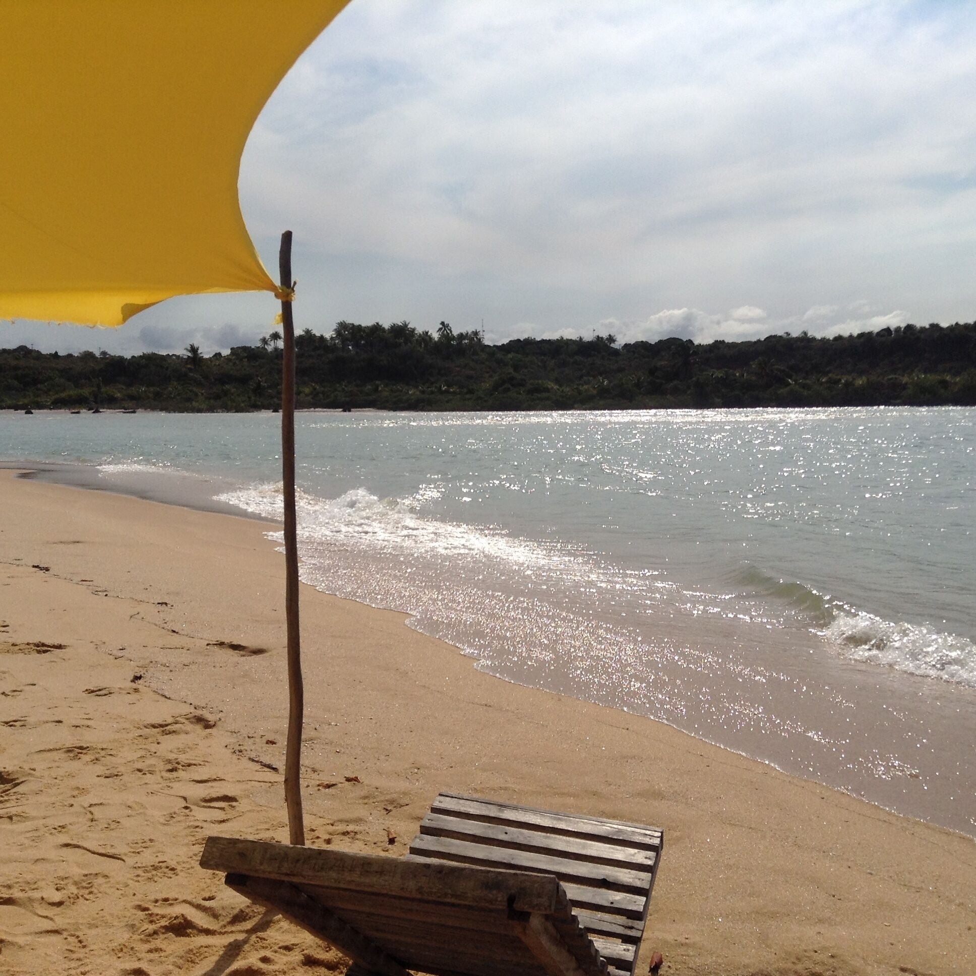 A beach with a wooden bench under a yellow umbrella, calm water, and a forested hill in the background.
