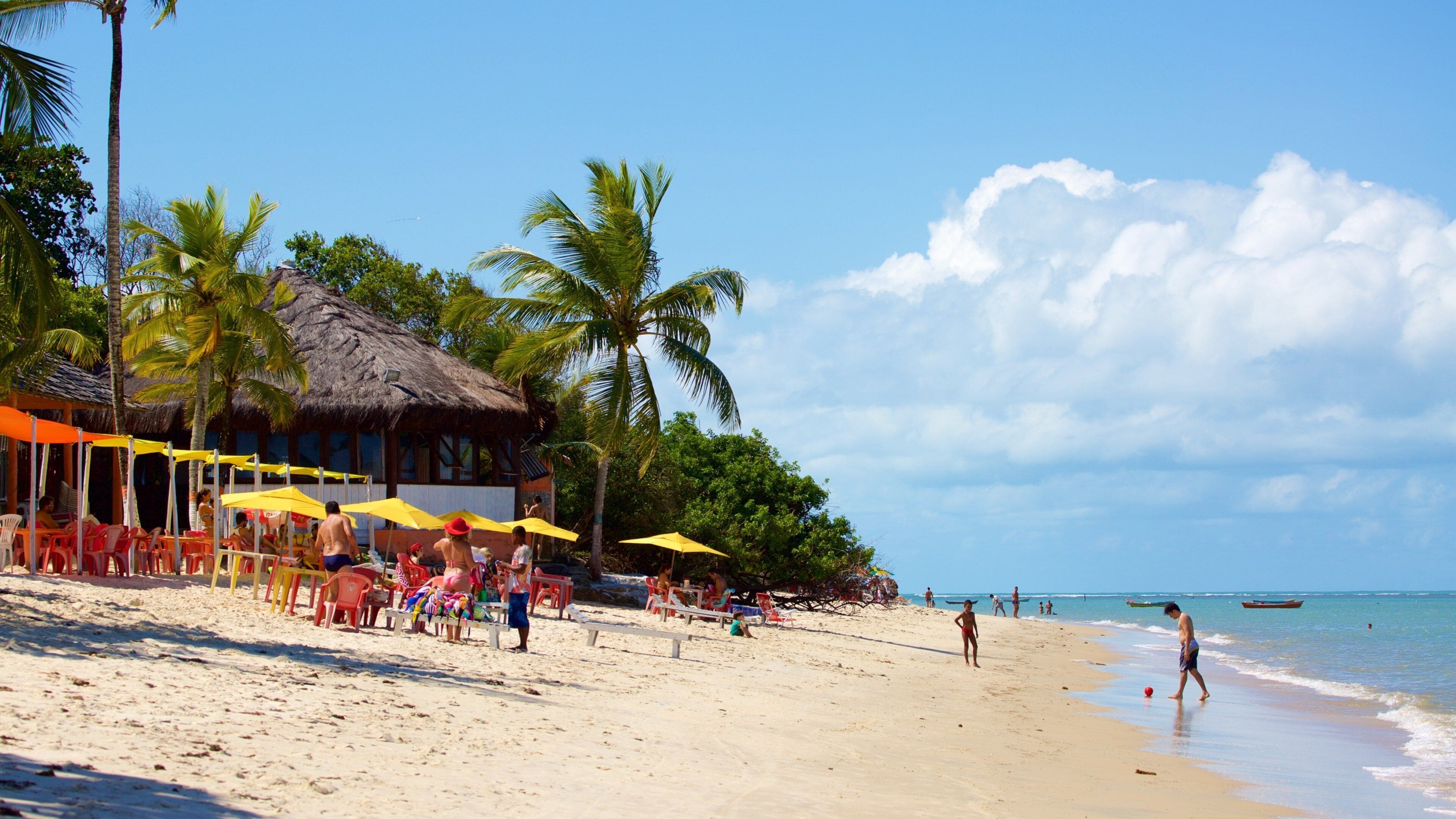 A beach with palm trees, a thatched-roof hut, and people relaxing under umbrellas.