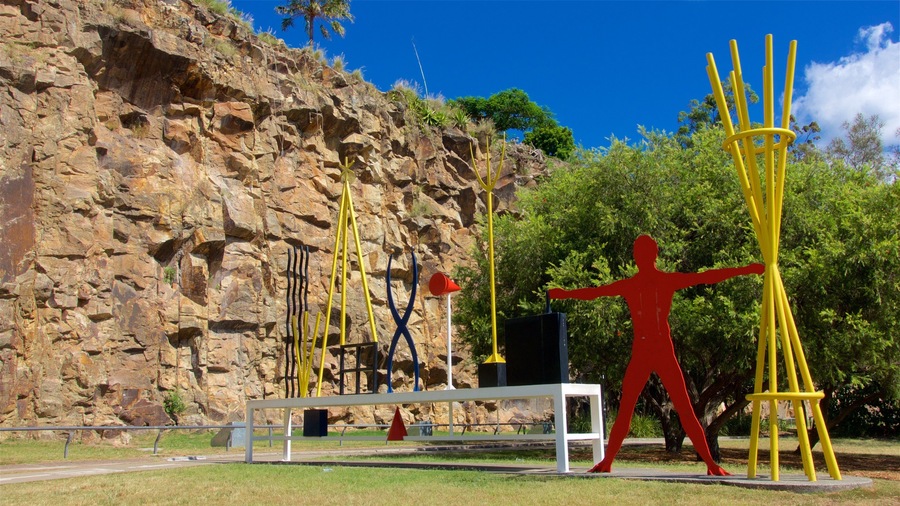 A sculpture of a human figure with outstretched arms in a park with a rocky backdrop.