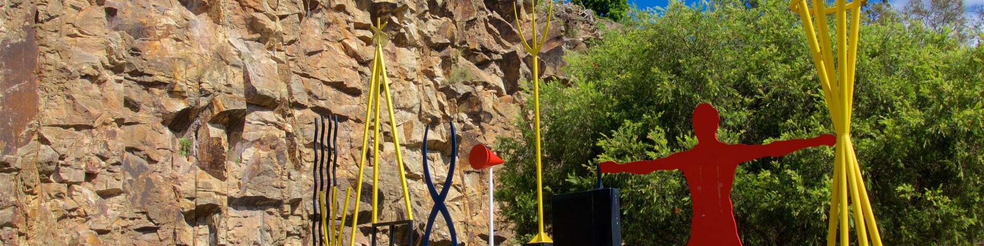Una escultura de una figura humana con los brazos extendidos en un parque con un fondo rocoso.