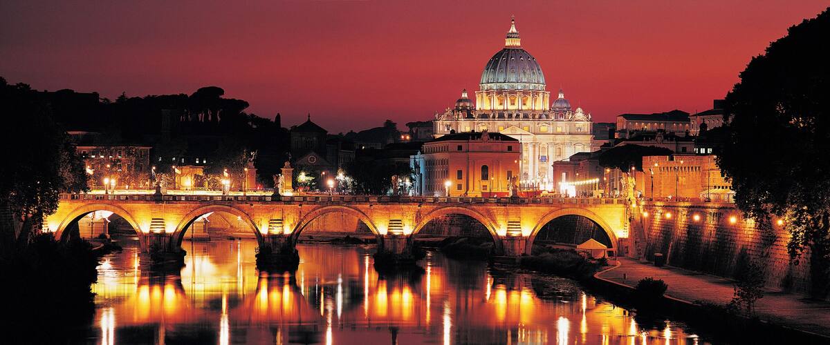 A lit bridge over a river with a large domed building in the background.