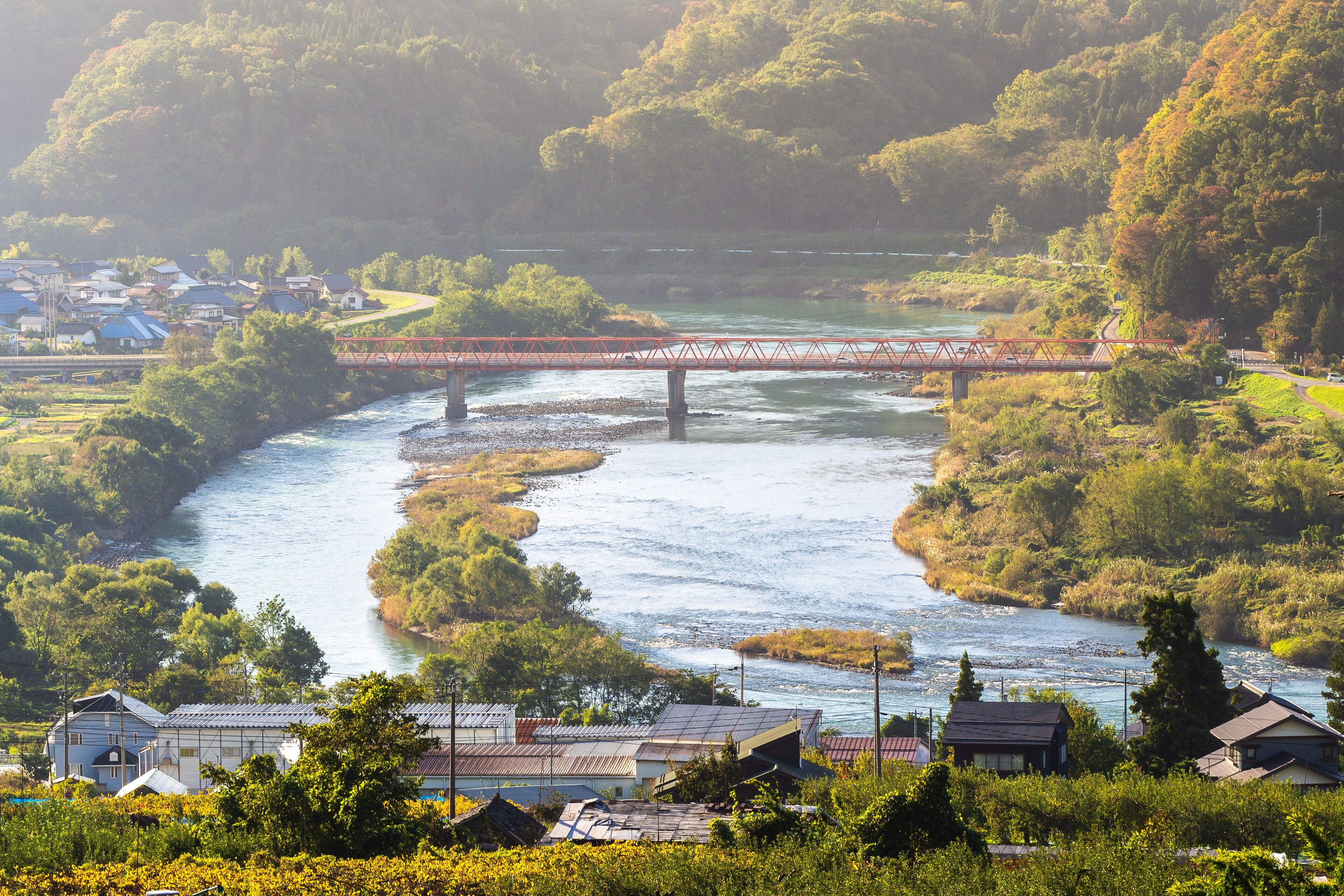 A river flowing through a valley with a bridge, houses, and lush greenery.