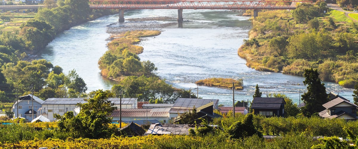 A river flowing through a valley with a bridge, houses, and lush greenery.