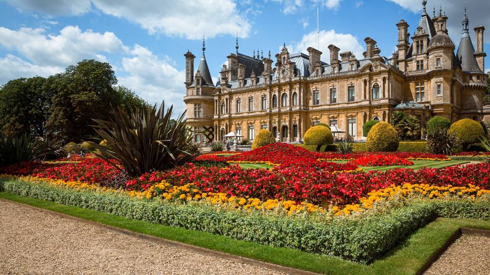 A grand historic building with ornate architecture, surrounded by meticulously landscaped gardens with vibrant flower beds.