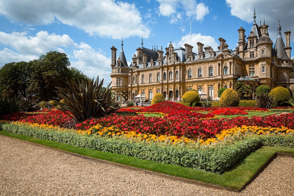 Un edificio histórico imponente con arquitectura ornamentada, rodeado de jardines meticulosamente cuidados y coloridos macizos de flores.