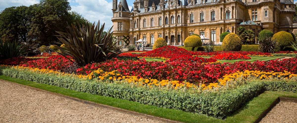 A grand historic building with ornate architecture, surrounded by meticulously landscaped gardens with vibrant flower beds.