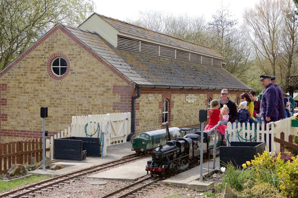 A small brick building with a white picket fence, a green steam train, and people near the train tracks.