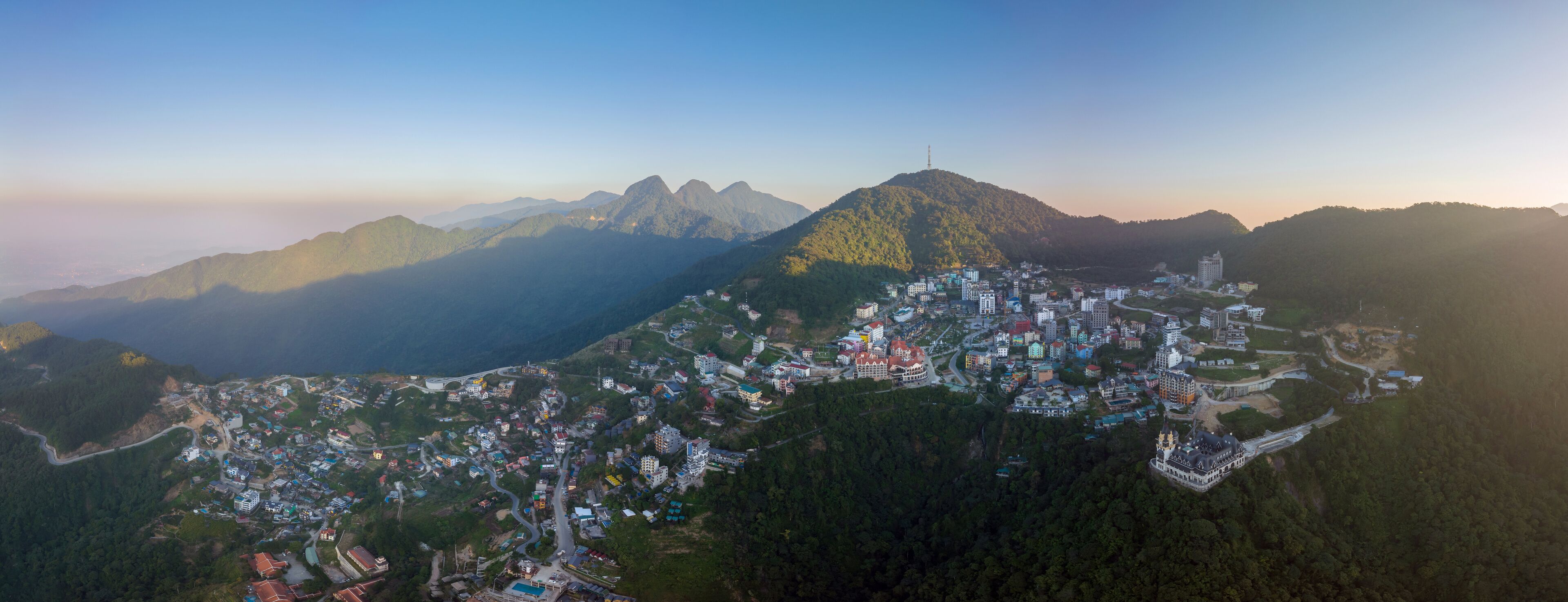 An aerial view of a mountainous town with dense housing and winding roads, surrounded by forested hills and distant peaks.