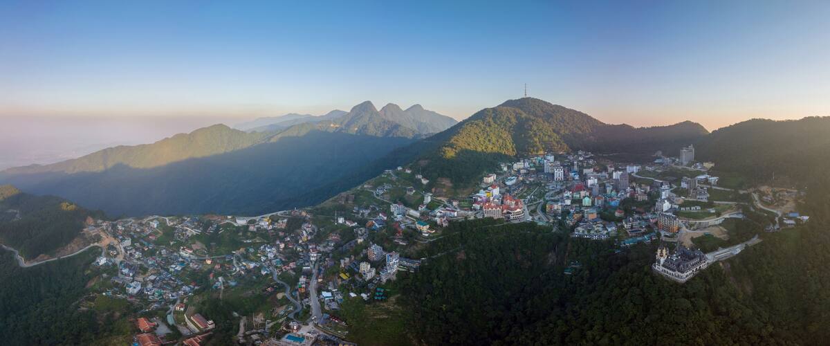 An aerial view of a mountainous town with dense housing and winding roads, surrounded by forested hills and distant peaks.