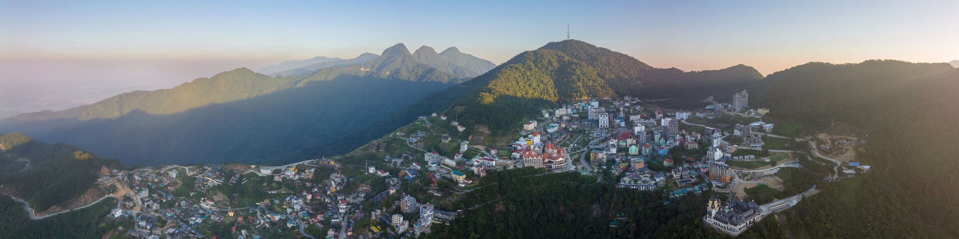An aerial view of a mountainous town with dense housing and winding roads, surrounded by forested hills and distant peaks.