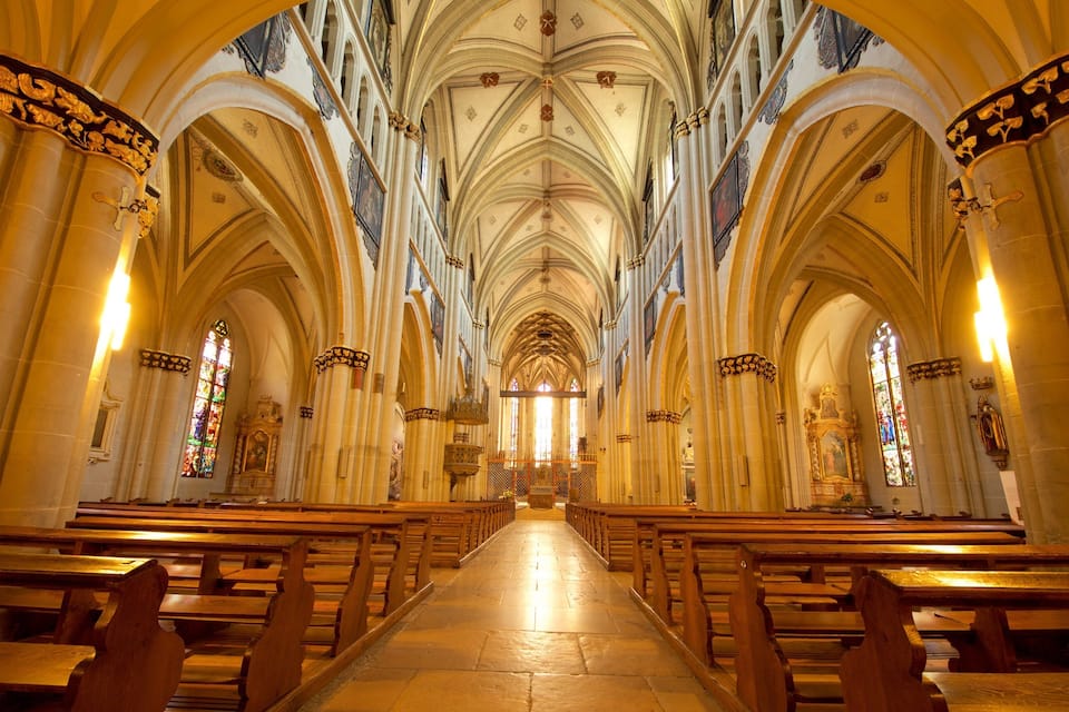 Interior de una catedral con altos techos abovedados, filas de bancas y vidrieras.