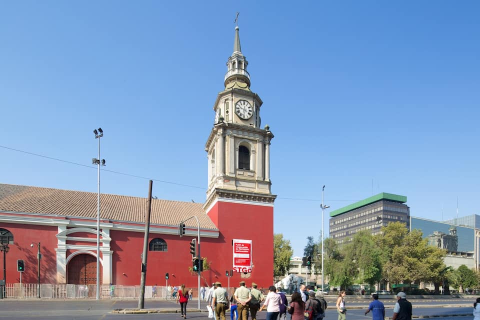 A historic building with a clock tower, a red facade, and a green-roofed modern building in the background.