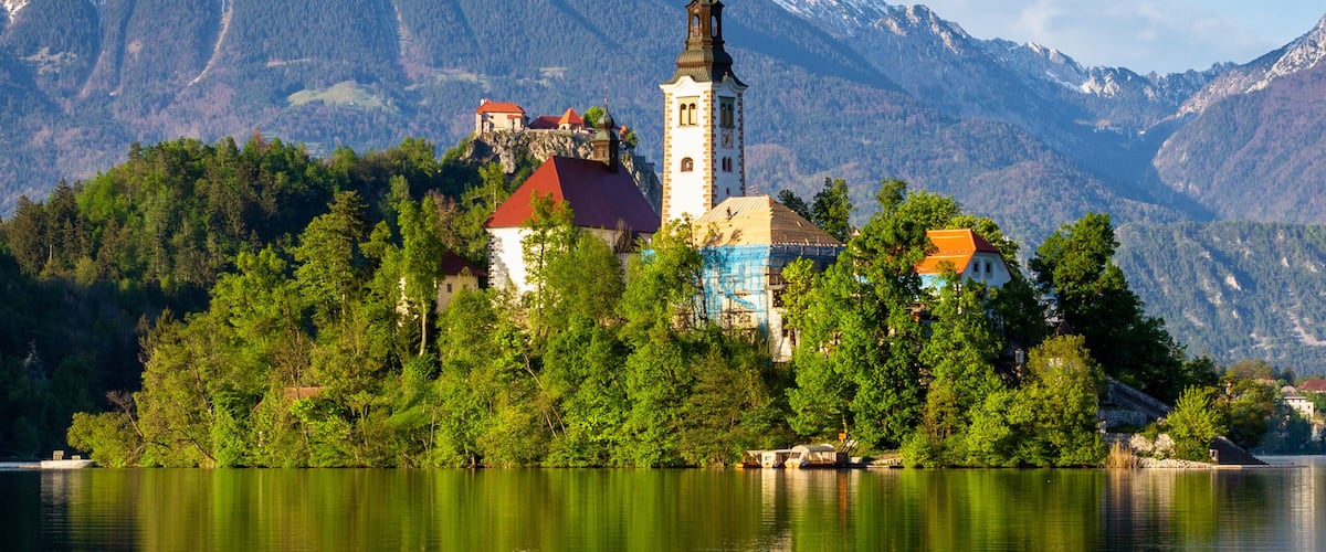 A church with a red roof on a lakeside island, surrounded by mountains and trees.