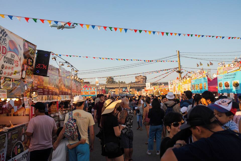 Um mercado de rua movimentado, com diversos quiosques de comida e decorações coloridas.