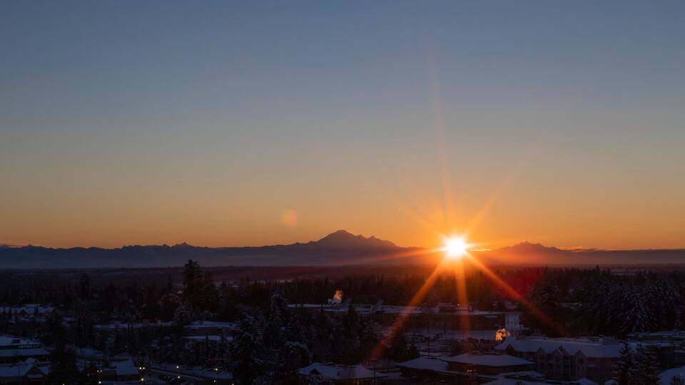 Sunburst over mountains and cityscape at sunset; Surrey, British Columbia, Canada
